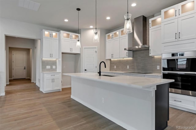 kitchen with white cabinets, sink, an island with sink, and wall chimney range hood