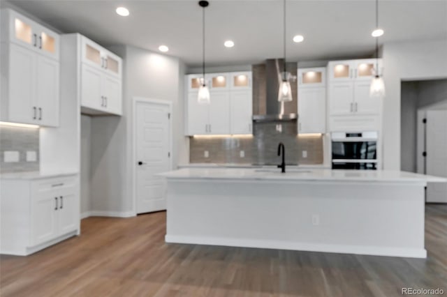 kitchen featuring wall chimney range hood, oven, an island with sink, and white cabinets