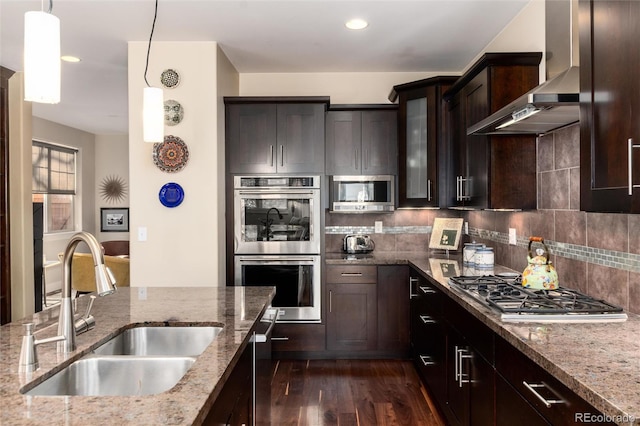 kitchen with dark brown cabinets, a sink, stainless steel appliances, wall chimney range hood, and dark wood-style floors