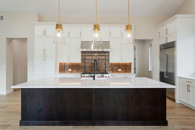 kitchen featuring a kitchen island with sink, stainless steel built in refrigerator, light stone counters, and range hood