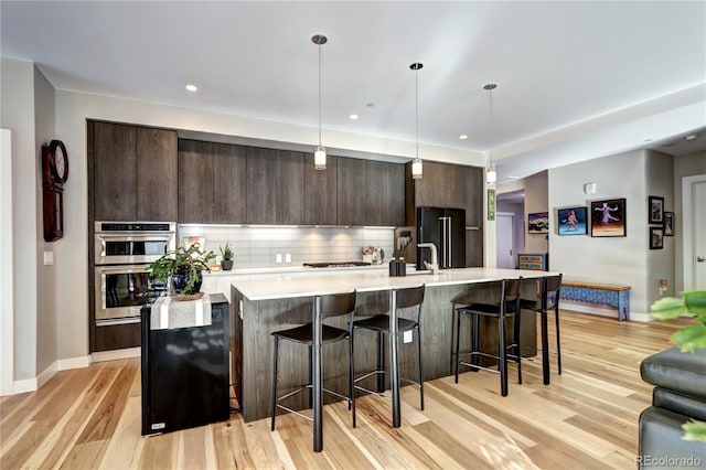 kitchen featuring dark brown cabinetry, hanging light fixtures, double oven, light hardwood / wood-style floors, and a center island with sink