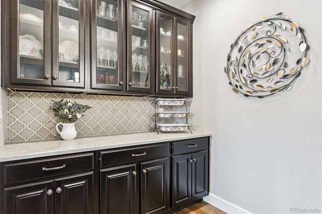 bar with light stone counters, backsplash, dark brown cabinetry, and light wood-type flooring