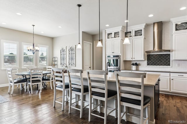 kitchen featuring wall chimney range hood, backsplash, hanging light fixtures, an island with sink, and white cabinets