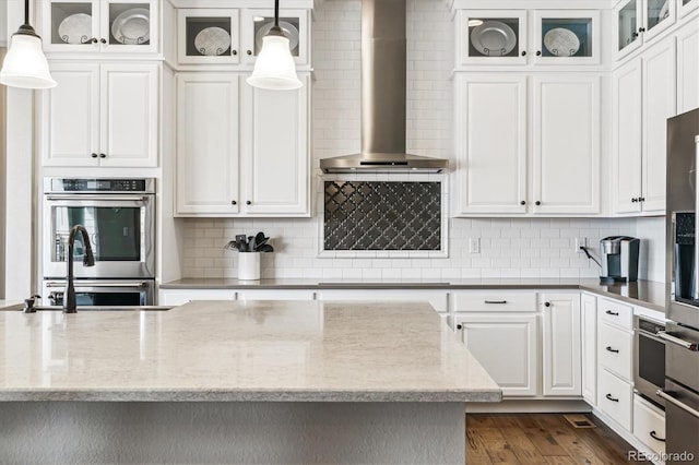 kitchen featuring pendant lighting, wall chimney range hood, white cabinetry, double oven, and light stone countertops