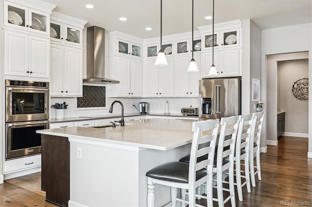 kitchen featuring a kitchen island with sink, light stone counters, wall chimney exhaust hood, and appliances with stainless steel finishes