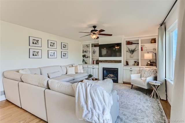 living area featuring built in features, a glass covered fireplace, ceiling fan, and wood finished floors