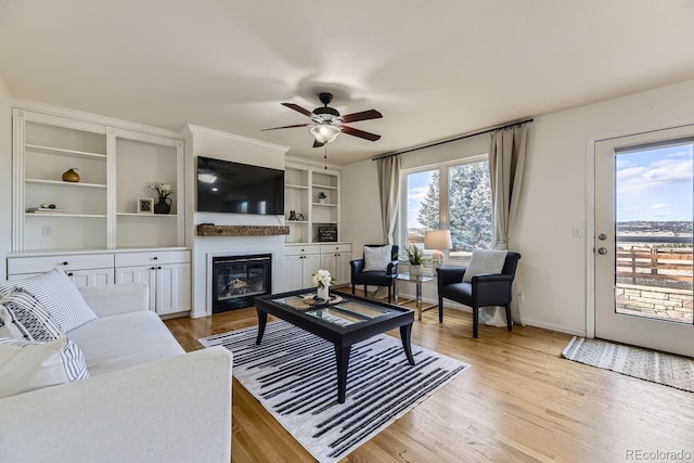 living area with a glass covered fireplace, ceiling fan, light wood-style flooring, and baseboards