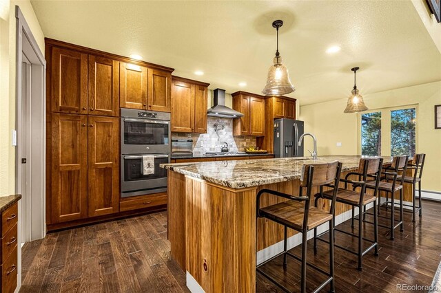459 County Road 85, Guffey, CO 80820 - kitchen featuring wall chimney range hood, stainless steel appliances, dark wood finished floors, brown cabinetry, and decorative backsplash