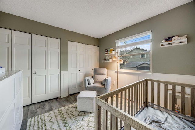 bedroom with a textured ceiling, wood finished floors, a wainscoted wall, a crib, and two closets
