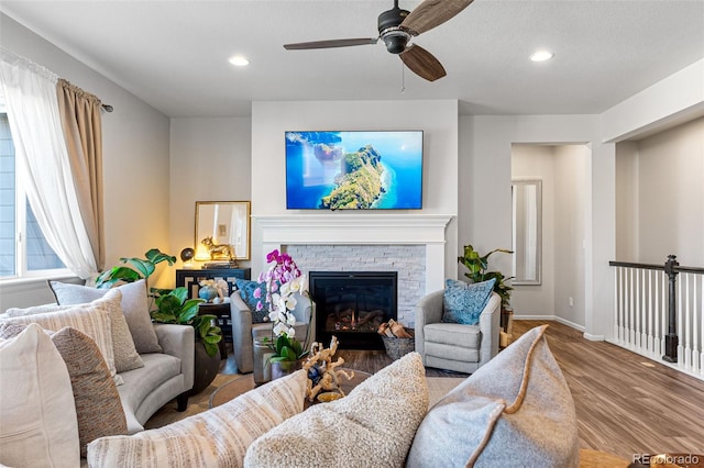 living room with ceiling fan, a stone fireplace, a textured ceiling, and light wood-type flooring