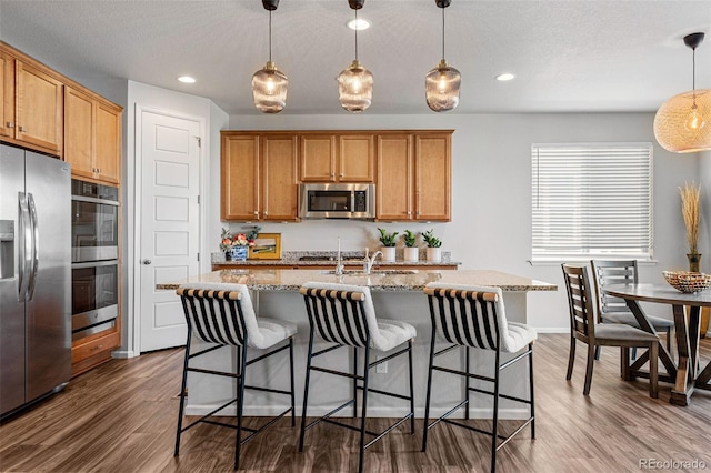 kitchen with pendant lighting, wood-type flooring, appliances with stainless steel finishes, and a kitchen island with sink