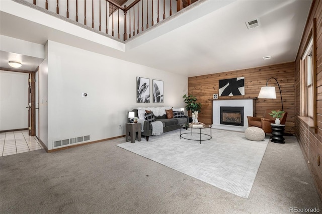 living room with wooden walls, carpet, visible vents, a high ceiling, and a fireplace with raised hearth