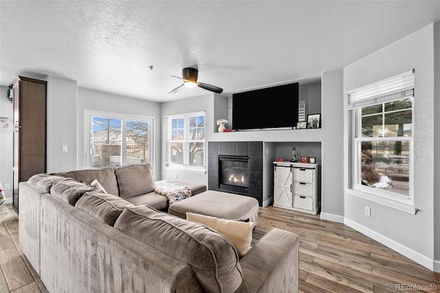 living area featuring ceiling fan, a textured ceiling, baseboards, light wood finished floors, and a tiled fireplace