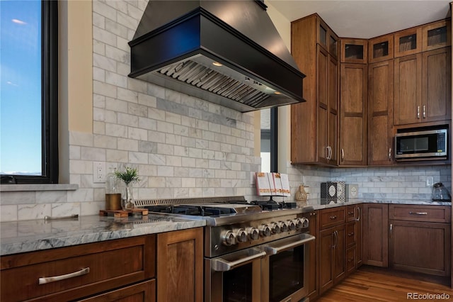 kitchen featuring light stone countertops, appliances with stainless steel finishes, tasteful backsplash, custom exhaust hood, and hardwood / wood-style floors