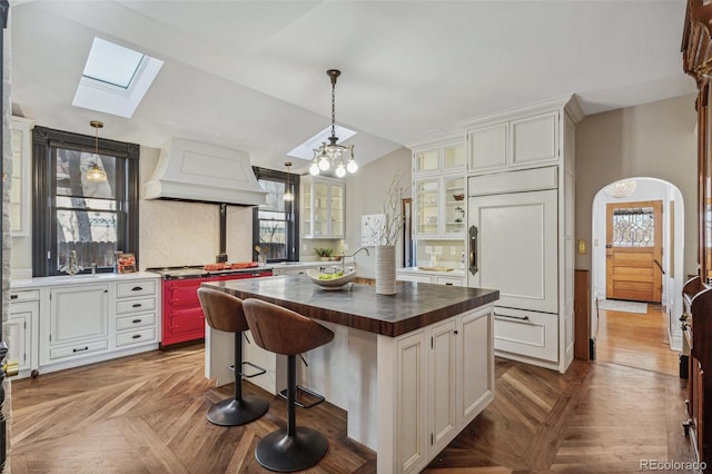 kitchen with vaulted ceiling with skylight, arched walkways, glass insert cabinets, premium range hood, and paneled fridge