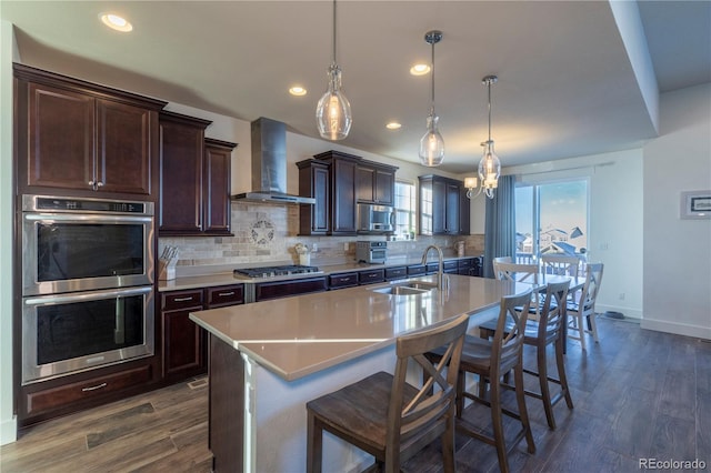 kitchen featuring appliances with stainless steel finishes, wall chimney exhaust hood, a kitchen island with sink, and hanging light fixtures