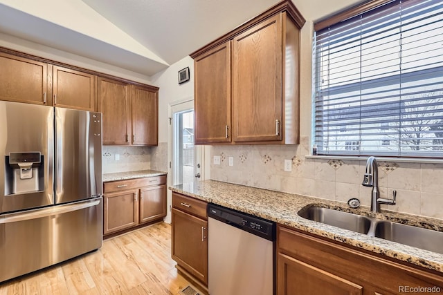 kitchen featuring lofted ceiling, light stone counters, stainless steel appliances, a sink, and brown cabinetry