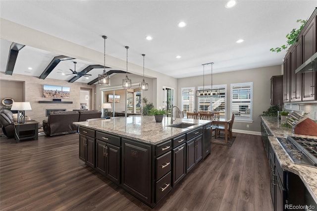 kitchen with decorative light fixtures, sink, light stone counters, dark brown cabinets, and a center island with sink