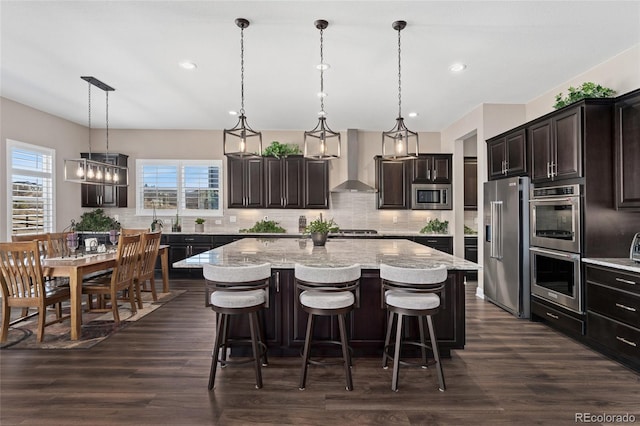 kitchen featuring a kitchen island, hanging light fixtures, stainless steel appliances, dark brown cabinets, and wall chimney exhaust hood