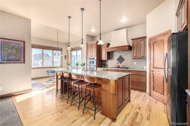 kitchen with appliances with stainless steel finishes, premium range hood, light stone counters, a center island with sink, and hanging light fixtures