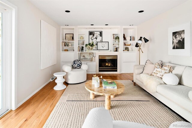 living area featuring light wood-style floors, built in shelves, baseboards, and a glass covered fireplace