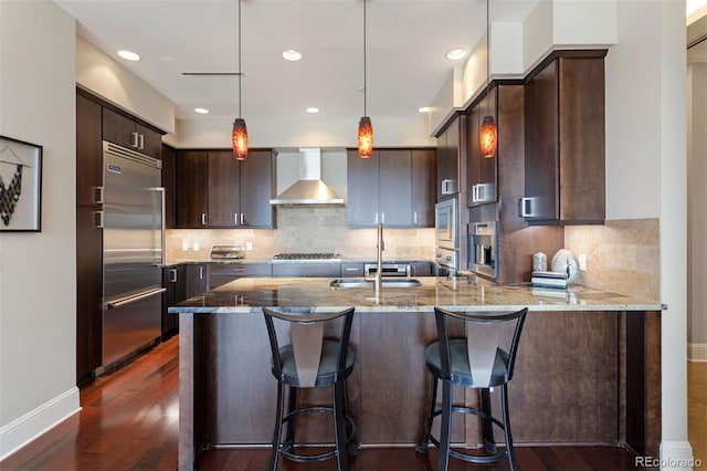 kitchen with kitchen peninsula, wall chimney exhaust hood, stainless steel appliances, light stone countertops, and dark hardwood / wood-style flooring