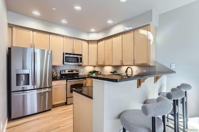 kitchen with light wood-type flooring, kitchen peninsula, stainless steel appliances, light brown cabinets, and a kitchen breakfast bar