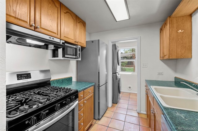 kitchen with stainless steel appliances, stacked washing machine and dryer, a sink, under cabinet range hood, and light tile patterned flooring