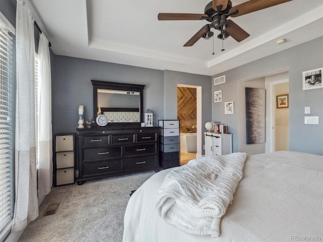 bedroom featuring a raised ceiling, light colored carpet, ensuite bath, and ceiling fan