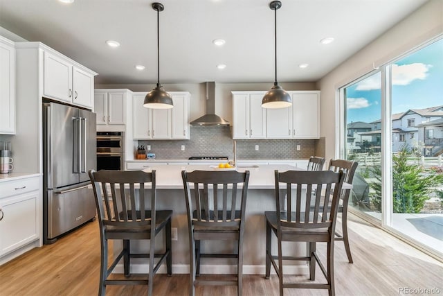 kitchen with appliances with stainless steel finishes, light wood-style floors, light countertops, wall chimney exhaust hood, and backsplash