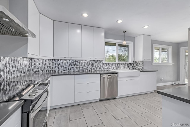 kitchen with appliances with stainless steel finishes, a sink, dark countertops, wall chimney range hood, and white cabinetry