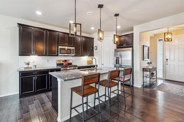 kitchen with appliances with stainless steel finishes, an island with sink, sink, dark stone countertops, and hanging light fixtures