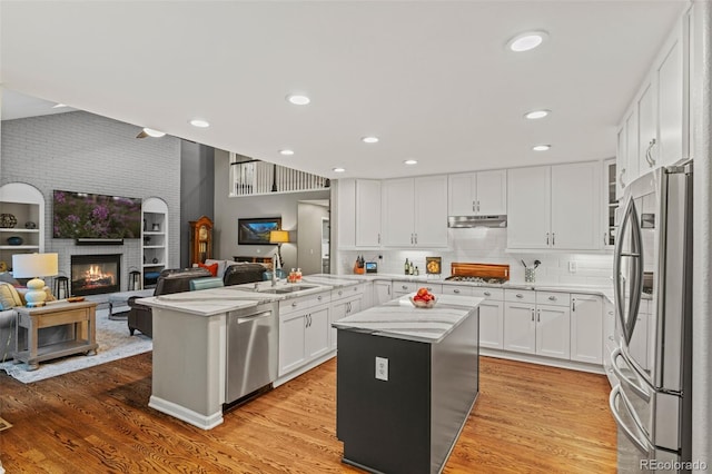 kitchen featuring light wood-style flooring, under cabinet range hood, white cabinetry, appliances with stainless steel finishes, and a brick fireplace