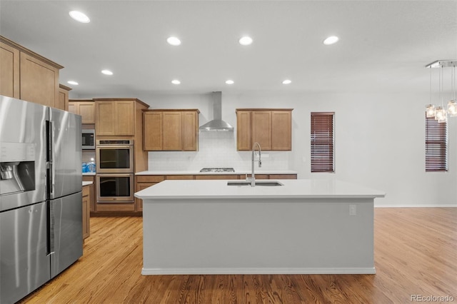 kitchen featuring sink, wall chimney exhaust hood, stainless steel appliances, decorative light fixtures, and a kitchen island with sink