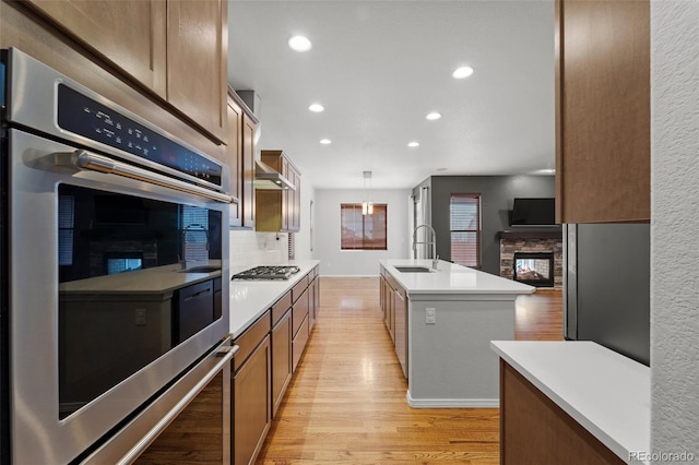 kitchen with pendant lighting, a kitchen island with sink, a stone fireplace, sink, and stainless steel appliances