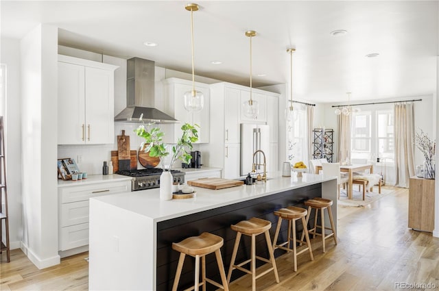 kitchen featuring white cabinetry, an island with sink, high quality fridge, decorative light fixtures, and wall chimney exhaust hood