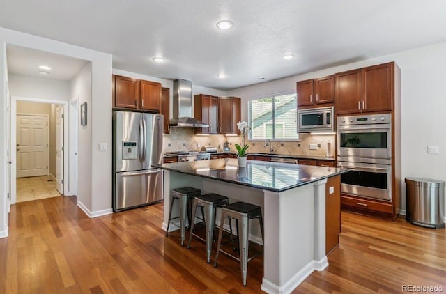 25402 E Glasgow Pl, Aurora, CO 80016 - kitchen featuring appliances with stainless steel finishes, wall chimney range hood, wood finished floors, a kitchen bar, and a center island