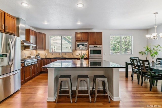 25402 E Glasgow Pl, Aurora, CO 80016 - kitchen with stainless steel appliances, wall chimney range hood, a kitchen bar, a chandelier, and decorative backsplash