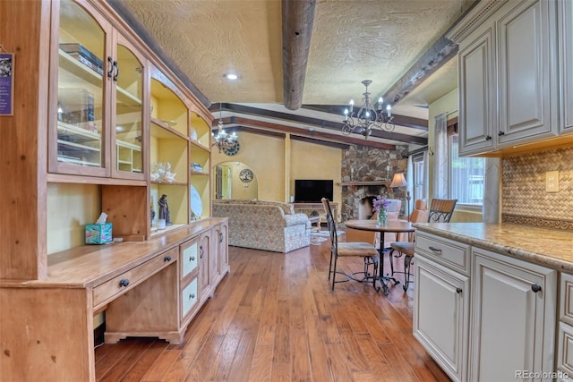 kitchen featuring a stone fireplace, decorative backsplash, light wood-type flooring, beamed ceiling, and a notable chandelier