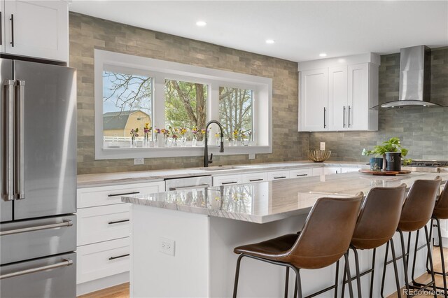 kitchen featuring high end refrigerator, wall chimney range hood, a sink, light stone countertops, and a kitchen island
