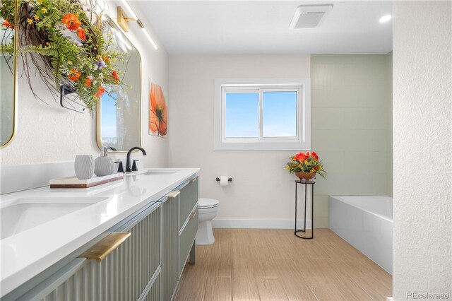 full bath featuring toilet, wood finished floors, double vanity, a tub to relax in, and baseboards