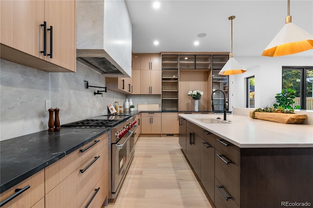 kitchen featuring high end stainless steel range oven, sink, hanging light fixtures, wall chimney exhaust hood, and light hardwood / wood-style flooring