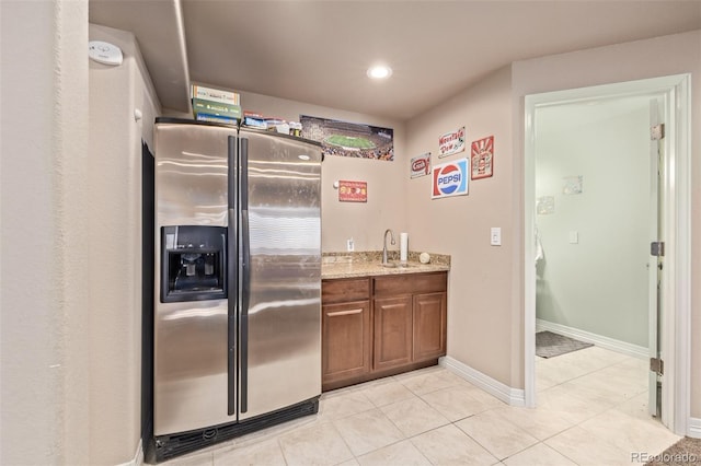 kitchen with light tile patterned floors, a sink, brown cabinetry, stainless steel refrigerator with ice dispenser, and light stone countertops