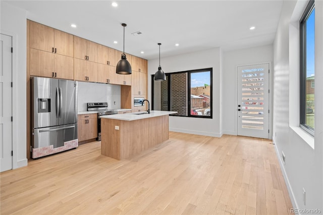 kitchen with light wood finished floors, hanging light fixtures, a kitchen island with sink, stainless steel appliances, and light countertops