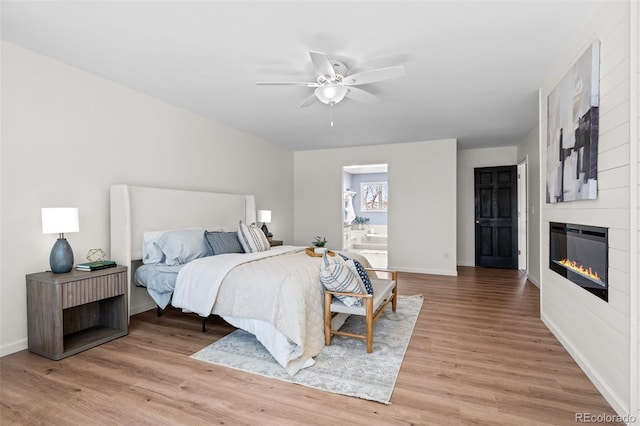 bedroom with light wood finished floors, ensuite bathroom, baseboards, a large fireplace, and ceiling fan