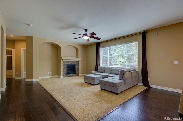living area featuring baseboards, a ceiling fan, dark wood-style flooring, and a stone fireplace