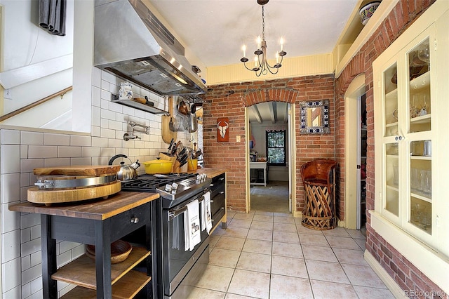 kitchen featuring stainless steel gas range oven, light tile patterned floors, an inviting chandelier, ventilation hood, and brick wall