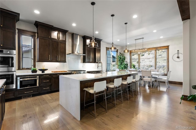 kitchen featuring wall chimney exhaust hood, light wood-style flooring, light countertops, stainless steel double oven, and a sink