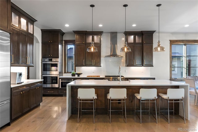 kitchen with light wood-style flooring, stainless steel appliances, light countertops, wall chimney range hood, and a center island with sink