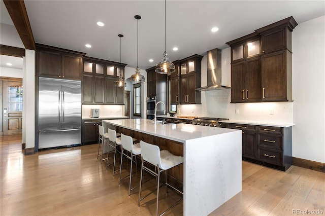 kitchen featuring light wood finished floors, backsplash, dark brown cabinetry, stainless steel built in refrigerator, and wall chimney exhaust hood